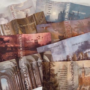 Peut inclure: Plusieurs couvertures de livres de fantasy, avec des titres tels que "Throne of Glass" et "Kingdom of Ash". Les couvertures présentent des illustrations de châteaux, de dragons et de paysages dans des tons sourds de bleu, de brun et de rouge.