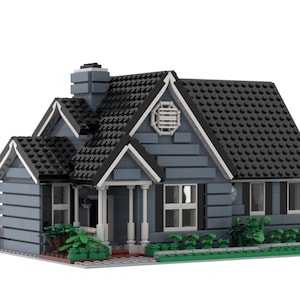 Op de afbeelding: Een gedetailleerd model van een huis gebouwd met in elkaar grijpende speelgoedblokken. Het huis heeft een grijze buitenkant, een zwart dak, witte afwerking en een kleine veranda. Groene struiken en een gazon zijn ook inbegrepen. Een kleinere afbeelding van het echte huis staat rechtsboven.