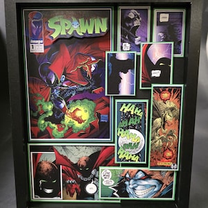 May include: Framed comic book art featuring Spawn. The main image is the cover of Spawn #1, with the title in green and red. Additional panels showcase comic book art with vibrant colors and action scenes, all within a black frame.