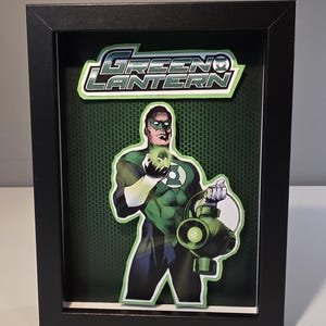 May include: A framed Green Lantern artwork. The image features the Green Lantern superhero in a green and black costume, holding a glowing green lantern and a green energy ball. The background is a textured green surface, and the frame is black.