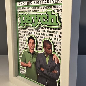 May include: Framed artwork featuring the word "Psych" in green, with two men in the center. The background includes text and the phrase "AND THIS IS MY PARTNER." The frame is white.