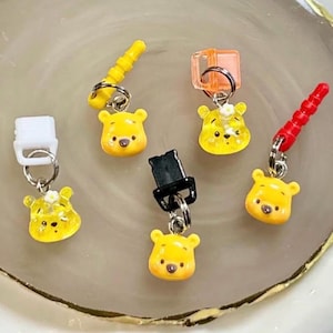 May include: A collection of yellow Winnie the Pooh-themed charms with various colored attachments. The charms are small, detailed, and feature the bear's face. Attachments include yellow, orange, red, white, and black pieces, all connected by silver rings.