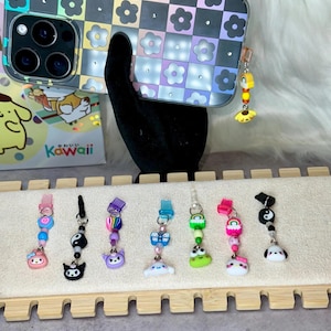 May include: A clear phone case with a checkerboard and flower design, held by a black hand. The case has a charm with a yellow sun and other colorful beads. Below are several phone charms with various cartoon characters.