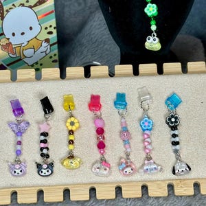 May include: A collection of colorful beaded charms with various character pendants. Charms include a butterfly, star, flower, and character designs. Each charm has a clip for attachment. The charms are displayed on a wooden rack.