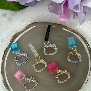 May include: A collection of colorful phone charms featuring a cat face design with sparkling rhinestones. The charms come in various colors, including blue, pink, and silver, and are attached to a phone jack.