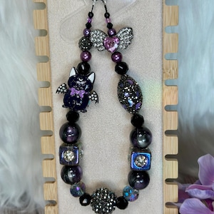 May include: A beaded keychain with a bat charm, heart-shaped pendant, and various purple and black beads. The bat charm has a purple bow tie. The keychain is attached to a silver-colored ring.
