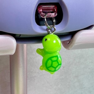 May include: A bright green turtle charm with a silver-colored ring attached to a pink plastic clasp. The turtle has a detailed shell design in a darker green. The background is a light purple and pink.