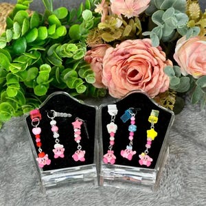 May include: Two pairs of dangle earrings displayed in clear acrylic cases. Each earring features a pink cartoon character charm, colorful beads, and star or flower accents. The background includes greenery and pink flowers.
