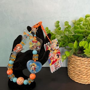 May include: A beaded keychain featuring orange, light blue, and striped round beads, a heart-shaped charm with a cartoon character, and a rectangular acrylic tag displaying a colorful anime-style illustration of multiple characters.