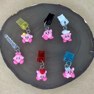 May include: A collection of pink Kirby-themed charms with various accessories, including a star, chef's hat, and heart. Each charm is attached to a silver-colored metal loop and a colored plastic plug. The charms are arranged on a gray, circular surface.