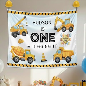 May include: A construction-themed birthday banner with yellow construction vehicles and the text "HUDSON IS ONE & DIGGING IT!". The banner has a light blue background with black and yellow hazard stripes.