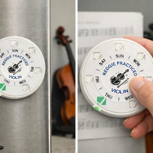 May include: White, circular magnet with the text "Reggie Practiced Violin" and a violin graphic. The days of the week are printed around the edge. A green check mark indicates the practice day. The magnet is on a refrigerator.
