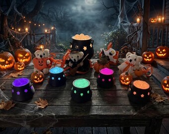 May include: A collection of black plastic cauldrons featuring star-shaped cutouts and glowing coloured contents, arranged on a wooden surface alongside plush teddy bears, small carved jack-o'-lanterns, and scattered autumn leaves.