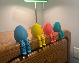 May include: Four colourful egg-shaped figurines with articulated legs sit on a wooden surface. The eggs are blue, yellow, coral, and teal. The figurines are under a green-shaded lamp, creating a whimsical and decorative display.