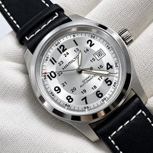 May include: A Hamilton Khaki Automatic watch with a silver dial, black Arabic numerals, and a date window at the 3 o'clock position. The watch features a stainless steel case and a black leather strap with white contrast stitching.