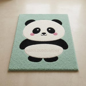 Könnte beinhalten: Ein rechteckiger getufteter Teppich mit hellminzgrünem Hintergrund und einer zentralen Pandabär-Grafik. Der Panda hat ein weißes Gesicht und einen weißen Bauch, schwarze Ohren, Gliedmaßen und Augenpartien sowie kleine rosa Wangen. Der Text ABDULQUDDUSRUGS ist sichtbar.