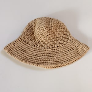 May include: A beige, crocheted bucket hat with a wide brim. The hat is made of a textured yarn, and the brim is slightly flared. The hat is a neutral color and would be suitable for sun protection.