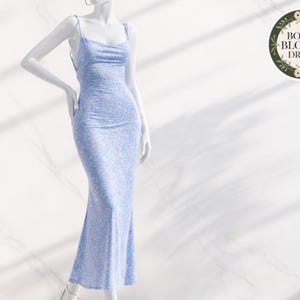 May include: A light blue floral print dress with a cowl neckline and spaghetti straps. The dress is displayed on a mannequin. The dress has a fitted silhouette that flares out at the hem. The dress is labeled "BOHO BLOOM DRESS".