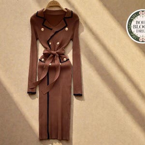 Op de afbeelding: Een bruine jurk met lange mouwen, een V-hals en een strikceintuur. De jurk heeft een dubbelborstig ontwerp met gouden knopen en zwarte bies. De jurk hangt aan een houten kleerhanger. De jurk is van de Boho Bloom Dress collectie.