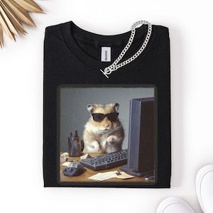 Może przedstawiać: Czarny t-shirt z zabawną grafiką przedstawiającą chomika w okularach przeciwsłonecznych pracującego przy komputerze. Obraz zawiera klawiaturę, mysz i monitor. Srebrny naszyjnik z łańcuszkiem jest zarzucony na koszulkę.