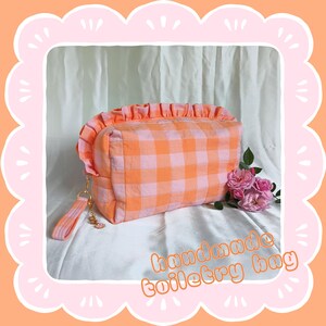 May include: A handmade toiletry bag in a gingham pattern of orange and pale pink. The bag features a ruffled top and a wrist strap. The text "handmade toiletry bag" is written in orange cursive. Pink roses are next to the bag.