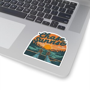 May include: A sticker with the words "Chase the Sunrise" in a retro style. The design shows a mountain landscape with a rising sun, and a person's legs and feet in the foreground. The sticker is placed on a laptop.