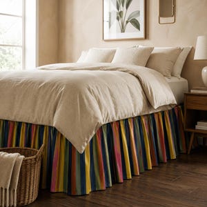 Op de afbeelding: Een bed met een beige dekbed en kussens, met een kleurrijke gestreepte bedrok in tinten blauw, geel, roze en groen. Een rieten mand met een beige deken staat aan het voeteneinde van het bed. De kamer heeft een neutraal kleurenpalet.