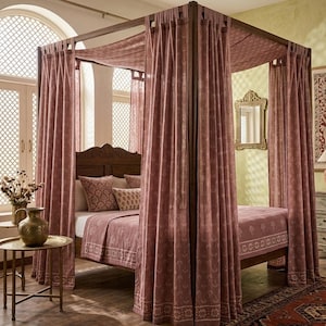 Puede incluir: Una cama con dosel con estructura de madera y cabecero ornamentado. La cama está cubierta con cortinas de color rosa polvoriento y ropa de cama a juego. Una pequeña mesa auxiliar con un jarrón decorativo está en primer plano y un espejo con marco dorado cuelga de la pared.