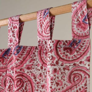 May include: Close-up of pink and white paisley patterned curtains. The curtains are hanging from a wooden rod. The fabric has a repeating design of swirling floral shapes in shades of pink and burgundy, with blue accents.