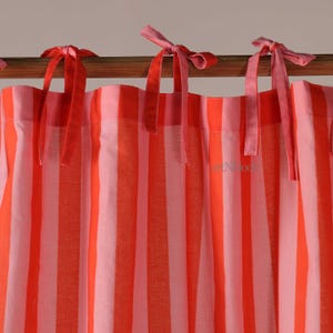 Può includere: Primo piano di una tenda a righe con strisce verticali alternate arancioni e rosa chiaro. La tenda è legata con nastri rosa. La tenda è appesa a un'asta di legno.