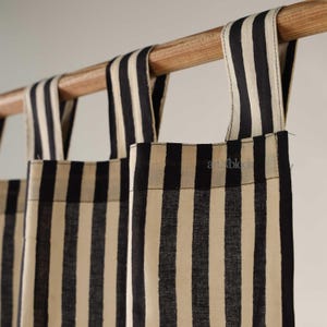 May include: Close-up of striped curtains in black and cream. The fabric is vertically striped and hangs from a wooden rod. The curtains have loops at the top for hanging. The text "art n black" is visible on the fabric.