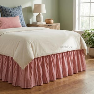 May include: A bed featuring a cream duvet cover and a red and white striped valance. Two pillows, one red and white striped and one blue patterned, are visible. A wooden bedside table with a lamp and decorative items sits beside the bed.
