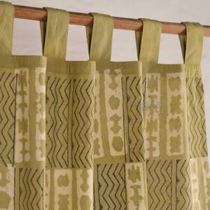 May include: Olive green curtains with a repeating geometric pattern in off-white and dark gray. The curtains are hanging from a wooden rod with matching olive green tabs at the top.