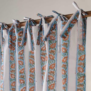 May include: White and blue striped curtains with orange floral accents. The curtains are tied to a wooden rod with white fabric ties. The design features a repeating floral pattern along the edges.