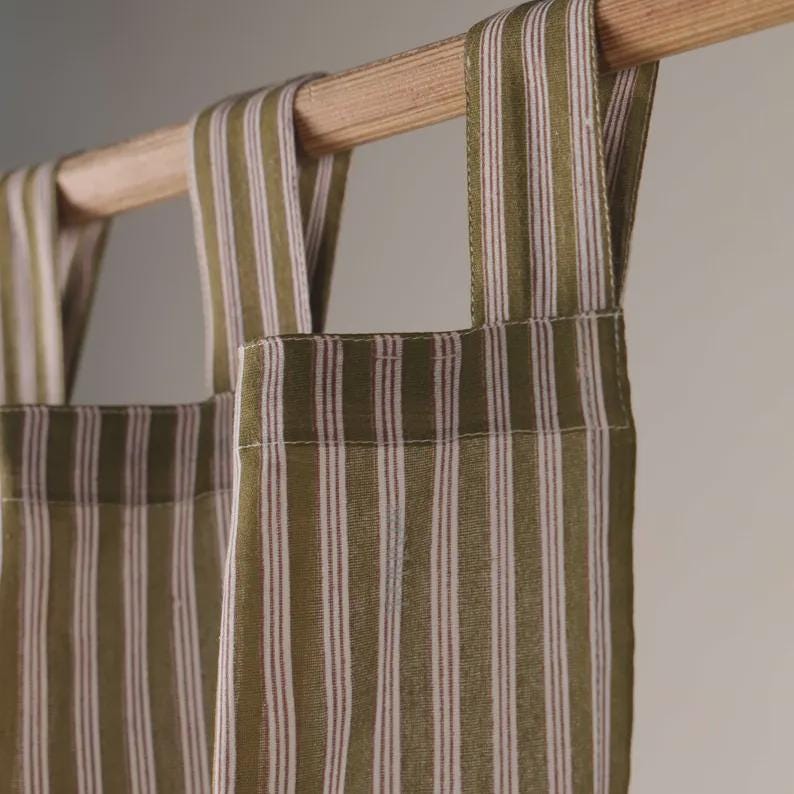 May include: Close-up of olive green and white striped curtains hanging from a wooden rod. The curtains have vertical stripes and are made of a soft fabric. The rod is a light brown color.