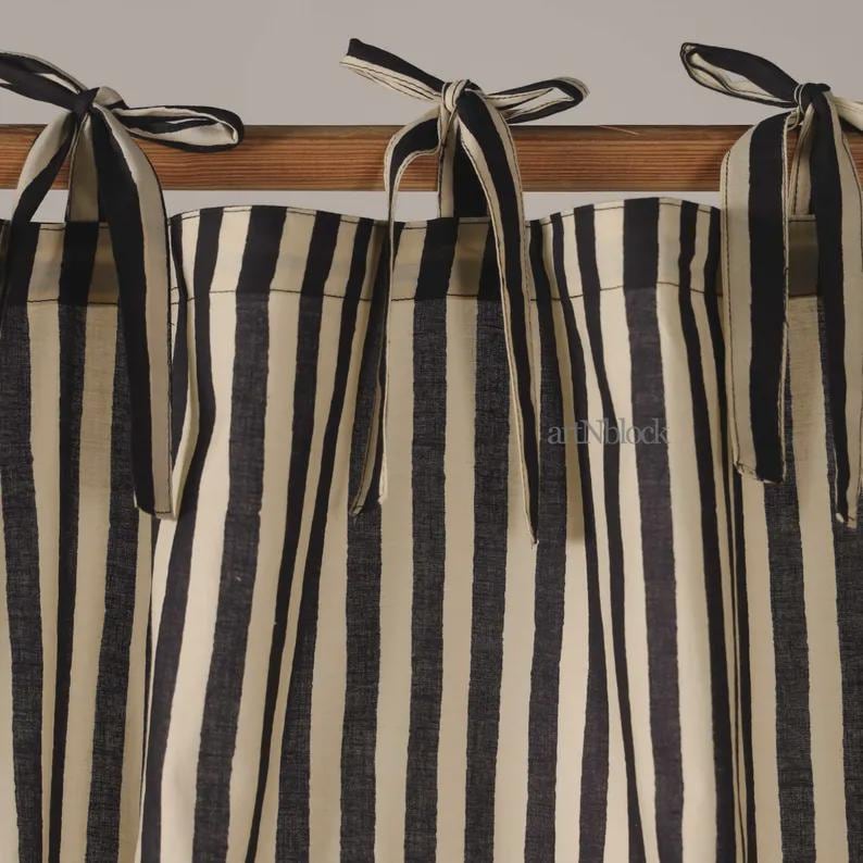 May include: Cream-colored and black striped curtains with matching tie-up closures. The fabric features vertical stripes, creating a classic and elegant look. The curtains are hanging on a wooden rod, adding a touch of warmth.