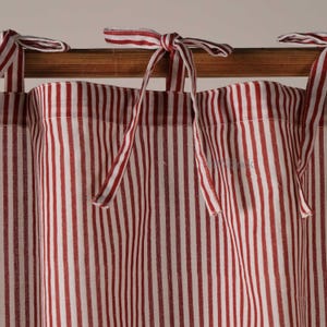 Puede incluir: Cortinas a rayas rojas y blancas con cintas para atar. La tela presenta rayas verticales, creando un diseño clásico. Las cortinas cuelgan de una barra de madera, añadiendo un toque rústico.