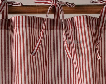 May include: A curtain panel featuring a vertical red and white striped pattern. The top edge of the fabric is attached to a wooden rod with multiple fabric ties that are knotted to secure the curtain in place, creating a gathered appearance.
