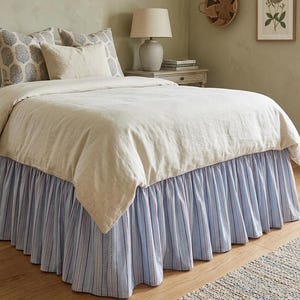 Pode incluir: Uma cama com edredão e almofadas de cor clara. A saia da cama é listrada em azul e branco com detalhes em vermelho. O quarto tem uma paleta de cores neutras com uma lâmpada e obras de arte.