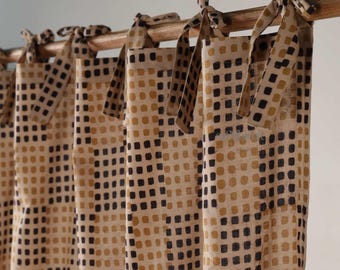 May include: Beige and brown patterned curtains with a repeating square design. The curtains are tied to a wooden rod with matching fabric ties. The fabric has a natural, textured appearance.