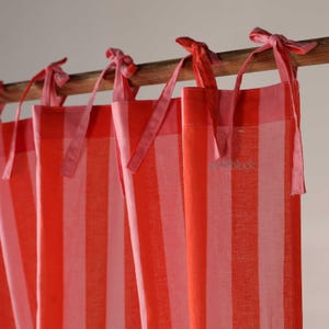 Puede incluir: Cortinas a rayas rojas y blancas con lazos rosas. Las cortinas cuelgan de una barra de madera. La tela tiene un aspecto suave y fluido. La palabra "block" está impresa en la tela.
