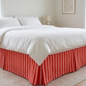 May include: A bed with white bedding and a red and pink striped bed skirt. The bed is in a well-lit room with a neutral color palette. The bed skirt has vertical stripes of varying widths.