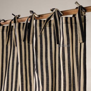 Puede incluir: Cortinas color crema con rayas verticales azul marino atadas a una barra de madera con cintas azul marino y crema. Las cortinas son de tela texturizada y cuelgan en suaves pliegues.