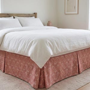 May include: A bed with a wooden headboard, white bedding, and a dusty rose bed skirt. The bed skirt features a repeating white floral pattern. The room has a neutral color palette with a vase on the nightstand.