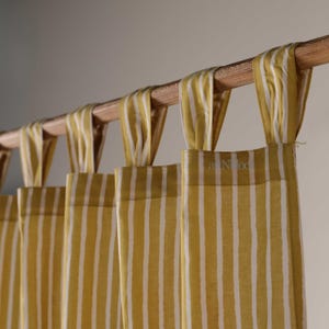 May include: Close-up of yellow and white striped curtains hanging from a wooden rod. The curtains are made of fabric and have vertical stripes. The rod is a light brown color. The curtains are hanging in front of a neutral-colored wall.