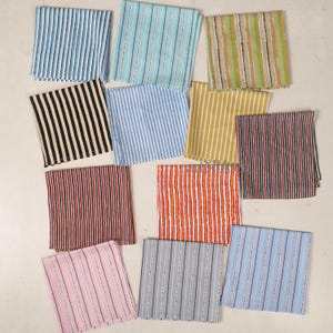 May include: A collection of folded fabric napkins featuring vertical stripes in various colours. The patterns include blue, black, red, orange, pink, grey, and yellow. Some stripes have thin accent lines. The napkins are arranged on a light surface.