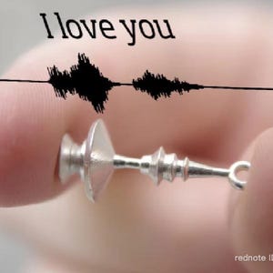 Op de afbeelding: Een kleine, zilverkleurige hanger met een uniek ontwerp wordt tussen vingers gehouden. Boven de hanger staan de woorden "I love you" in het zwart, vergezeld van een zwarte golfvormige grafiek. De hanger heeft een cilindrisch lichaam met een lus.