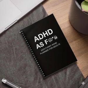 以下が含まれることがあります： 白字で「ADHD AS F@*#」と書かれた黒いリング綴じプランナー。下には「2025 FUNNY ADHD PLANNER FOR ADULTS」と書かれています。プランナーは濃い灰色の表面にあり、ラップトップとペンが見えます。