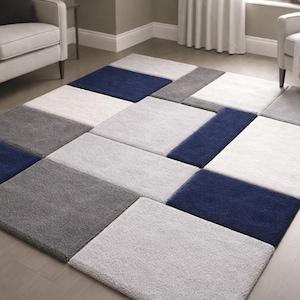 Könnte beinhalten: Rechteckiger Teppich mit geometrischem Muster. Der Teppich besteht aus rechteckigen Blöcken in Weiß, Grau und Marineblau. Er liegt auf einem hellen Holzboden. Im Hintergrund sind ein Sofa und ein Sessel zu sehen.