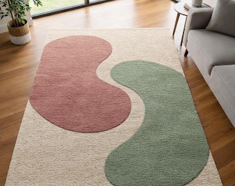 May include: A rectangular area rug with a cream-colored background and two large, abstract shapes in dusty rose and sage green. The rug is made of a soft, textured material, suitable for a living room or bedroom.